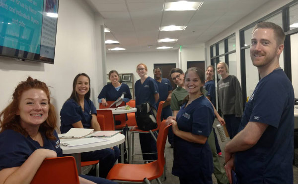 A group of students in scrubs sitting and standing around tables with orange chairs